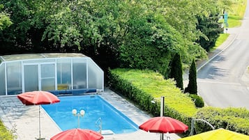A swimming pool with red and yellow umbrellas, surrounded by lush greenery and a road in the background.