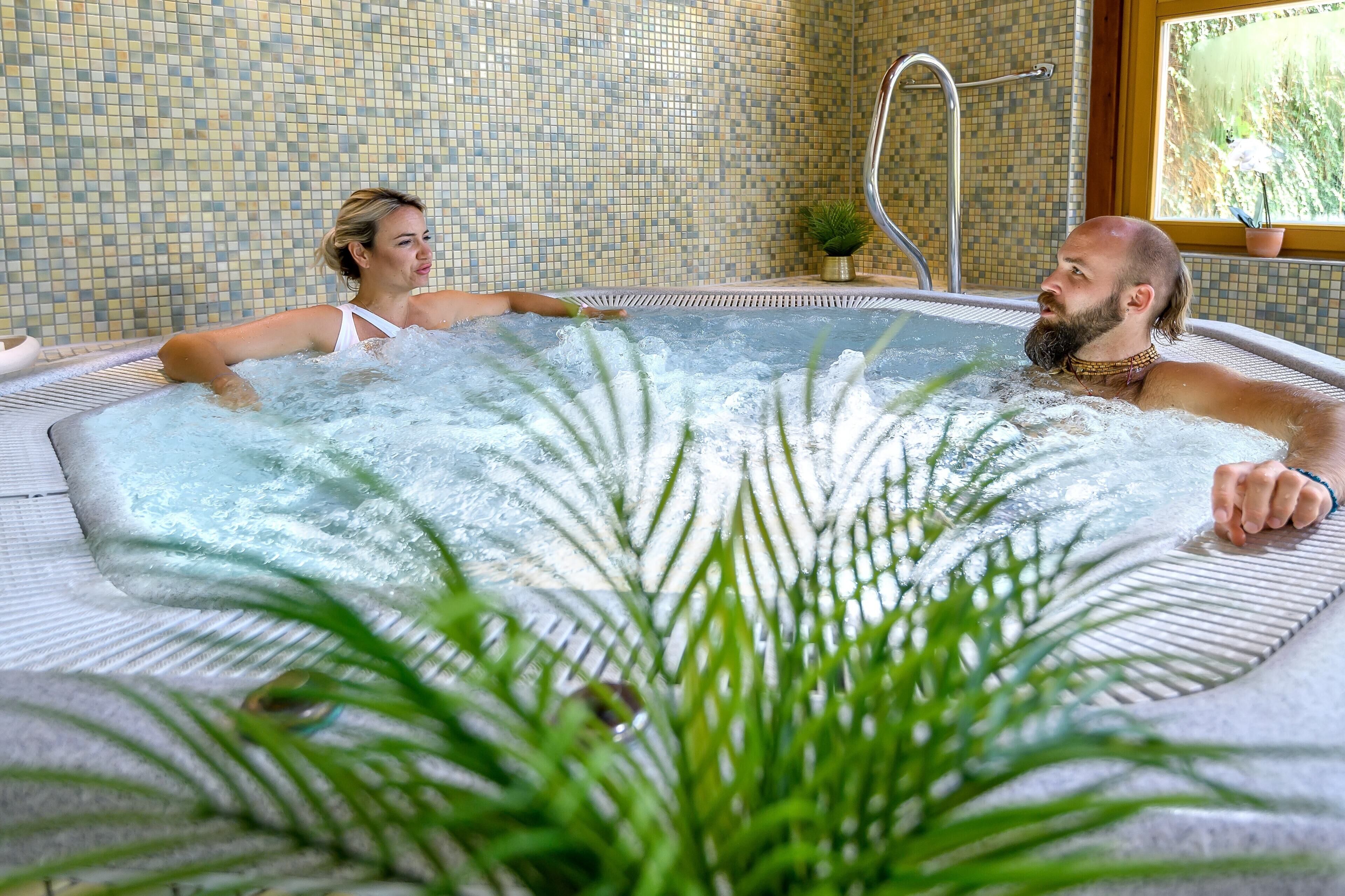 A hot tub with two people, surrounded by a tiled wall and a potted plant.