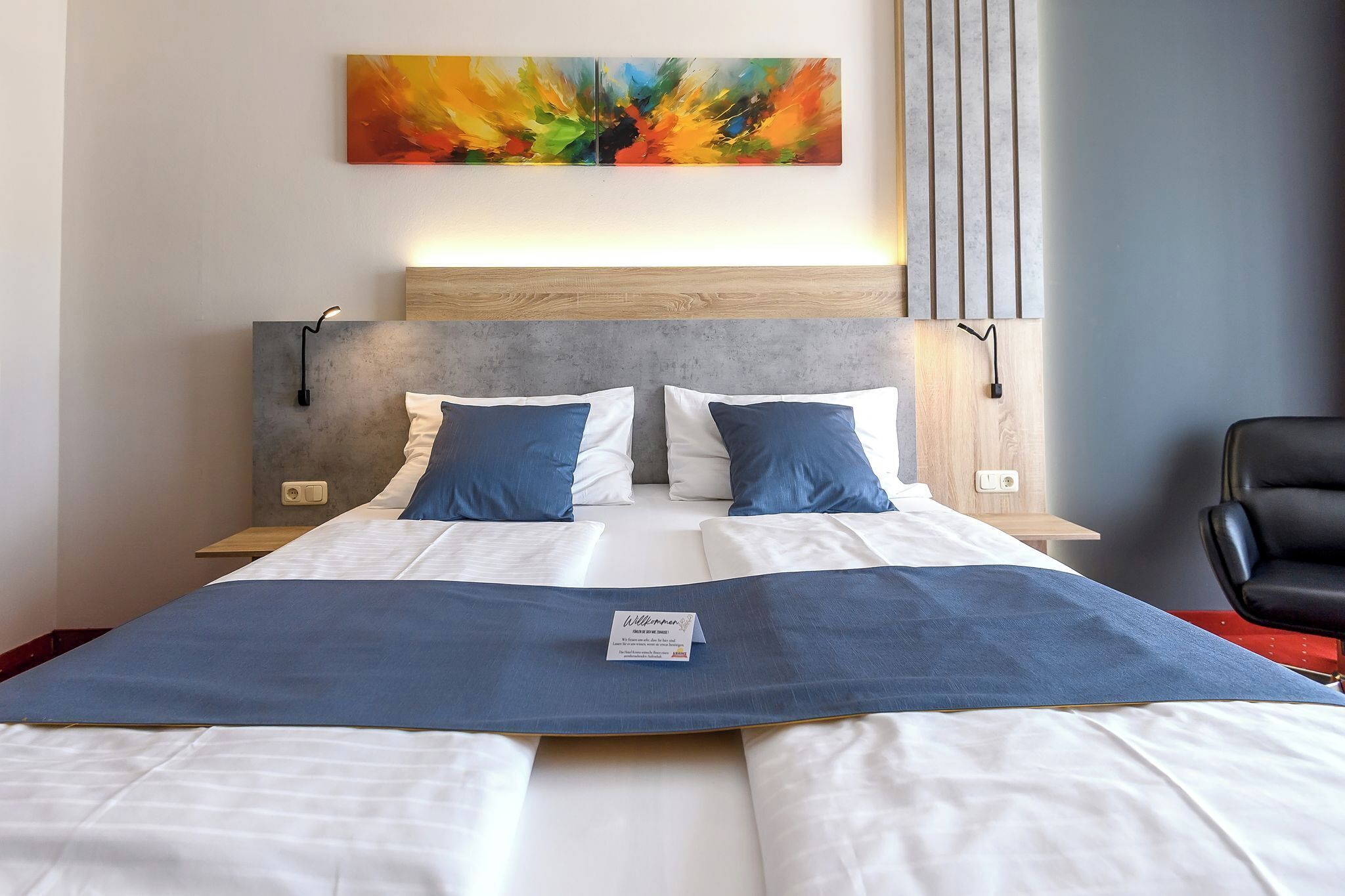 Ein ordentlich bezogenes Doppelbett mit blauem Akzent, ein Holz-Kopfteil und ein farbenfrohes, abstraktes Gemälde darüber.