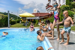 A swimming pool area with people, a building with a red roof, and a yellow umbrella. - Vital Hotel Krainz mit Nebenhaus Familien Hotel (Jennersdorf)