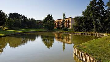 A tranquil pond with a wooden fence, surrounded by lush greenery and a multi-story building in the background.