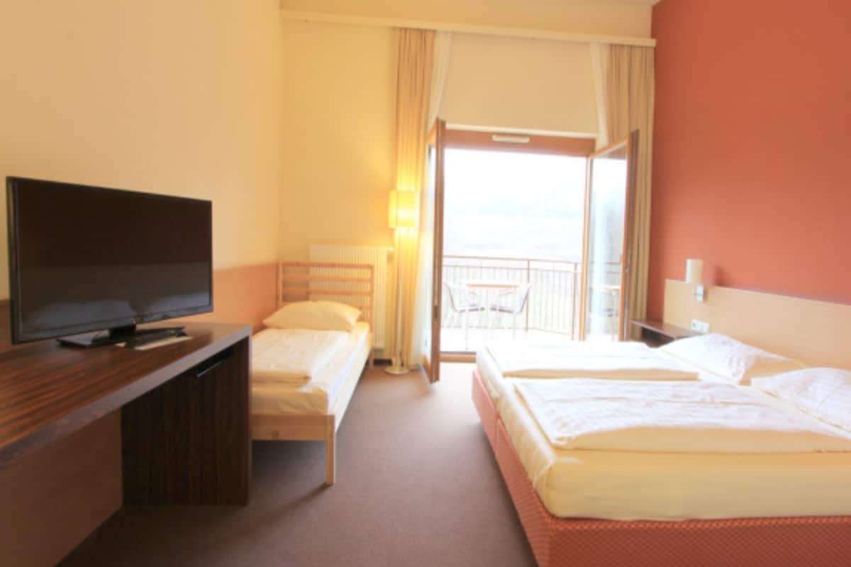 A hotel room with two beds, a TV on a wooden desk, a balcony with a view, and a window with curtains.