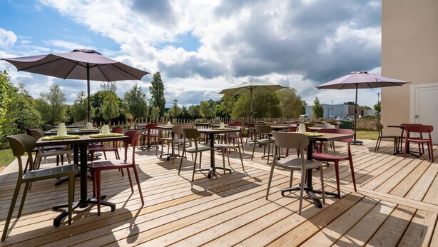 Eine Terrassengaststätte mit mehreren Tischen und Stühlen, beschattet durch Sonnenschirme, umgeben von Bäumen.