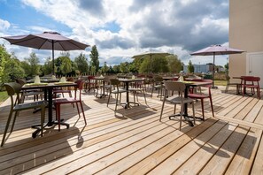 Une terrasse extérieure dotée de plusieurs tables et chaises, ombragée par des parasols et entourée d’arbres.