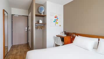 Habitación de hotel con cama, escritorio con silla, una pequeña estantería con un ventilador y una obra de arte colgada en la pared.