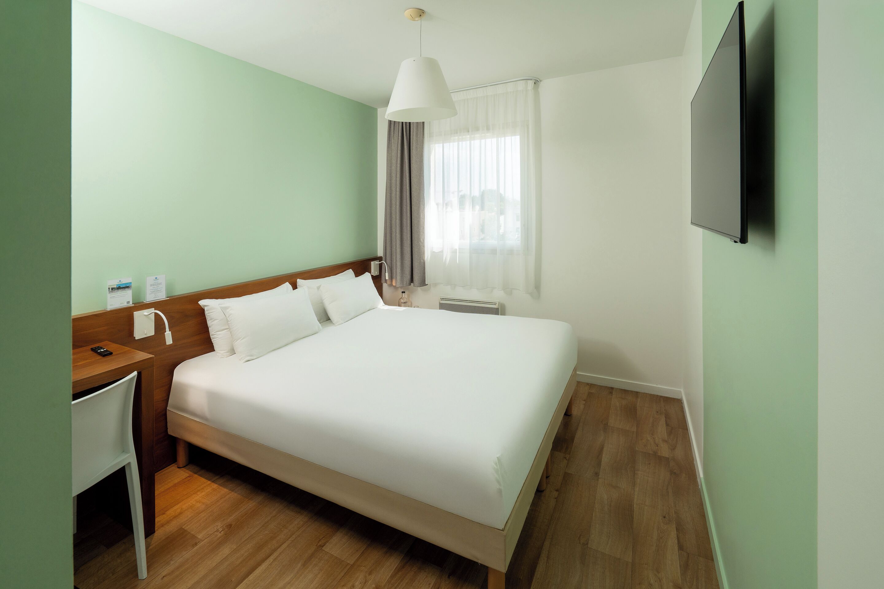 A neatly arranged hotel room with a bed, a desk, a chair, a TV, and a window with curtains.