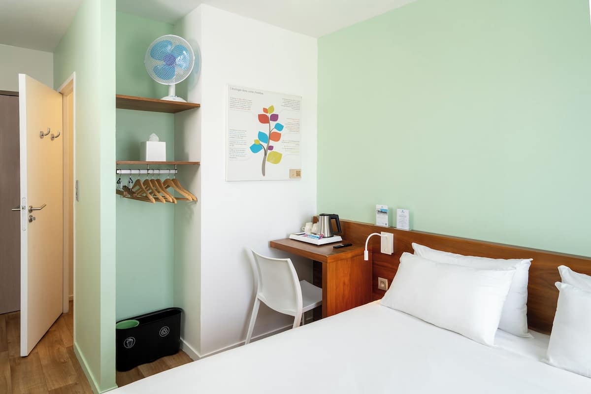 Une chambre d’hôtel équipée d’un lit, d’un bureau, d’une chaise, d’un ventilateur et d’une étagère fixée au mur, sur laquelle se trouvent une serviette et une boîte de mouchoirs.
