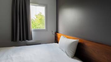 Una habitación de hotel con cama, ventana con cortinas, radiador y pared con acabado oscuro.
