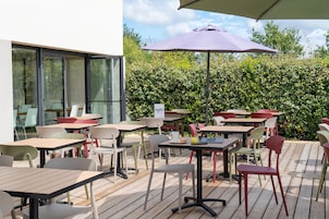 An outdoor café with tables and chairs, a purple umbrella, and a hedge in the background.