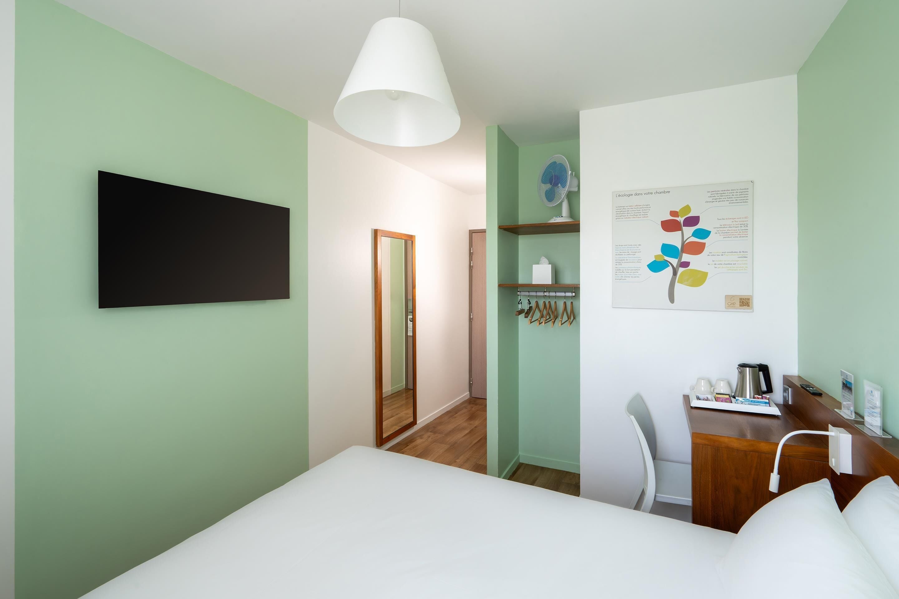 A hotel room with a bed, a desk, a chair, a mirror, a wall-mounted TV, and a poster.