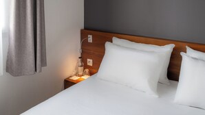 Ein ordentlich bezogenes Bett mit weißen Leinen, einem hölzernen Kopfteil und einem Nachttisch mit Flasche, Gläsern und einer kleinen Lampe.