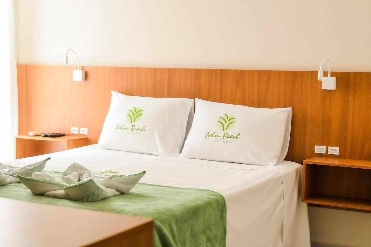 A hotel room with a wooden headboard, two pillows with 'Palm Beach' branding, a green bedspread, and a bedside table with a lamp.
