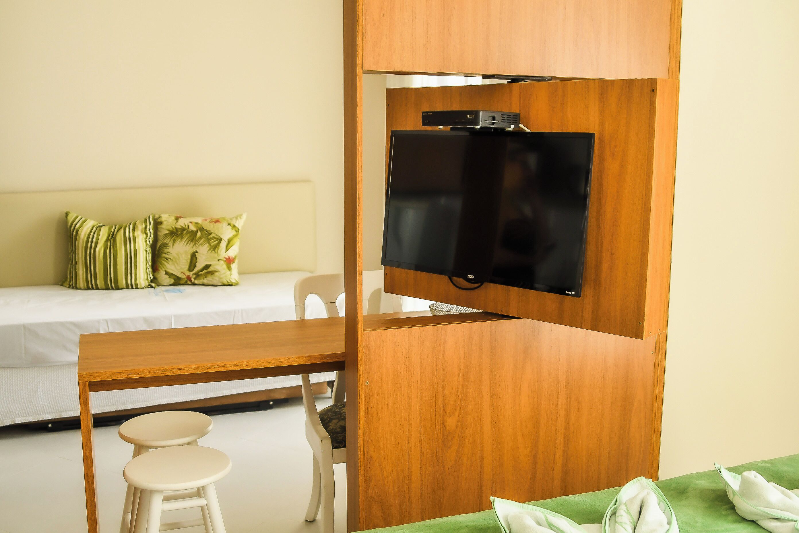 A room with a bed, a TV mounted on a wooden partition, a dining table with two stools, and a green couch.