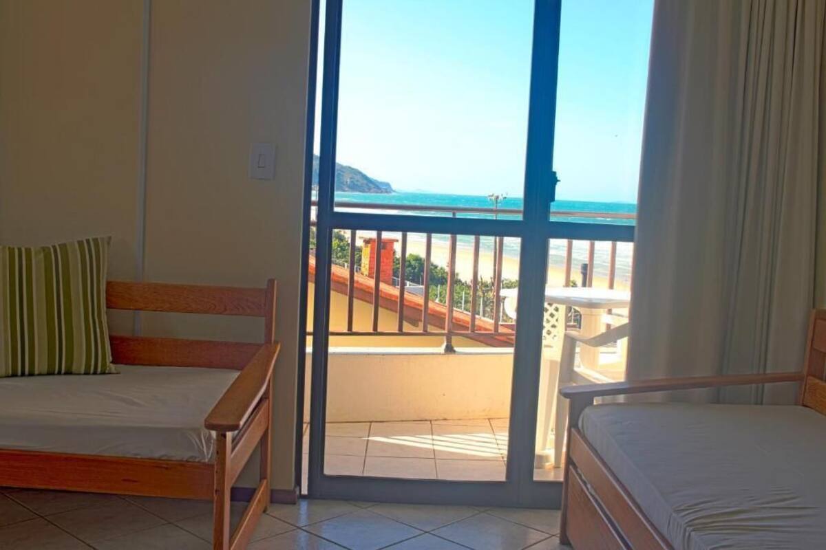 A room with two wooden beds, a sliding glass door leading to a balcony with a view of the beach and ocean.