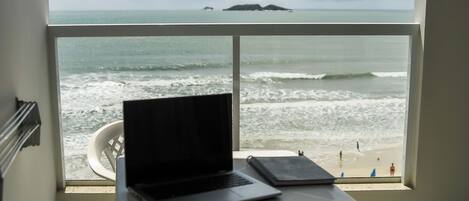 A laptop on a table by a window with a view of the ocean and beach.