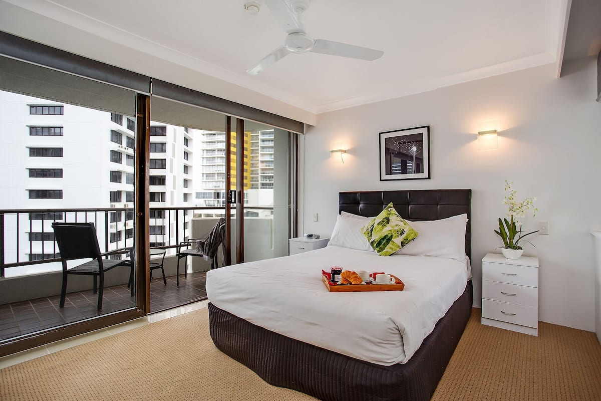 A hotel room with a bed, a nightstand, a wall-mounted light, a ceiling fan, a sliding glass door to a balcony, and a view of a building outside.