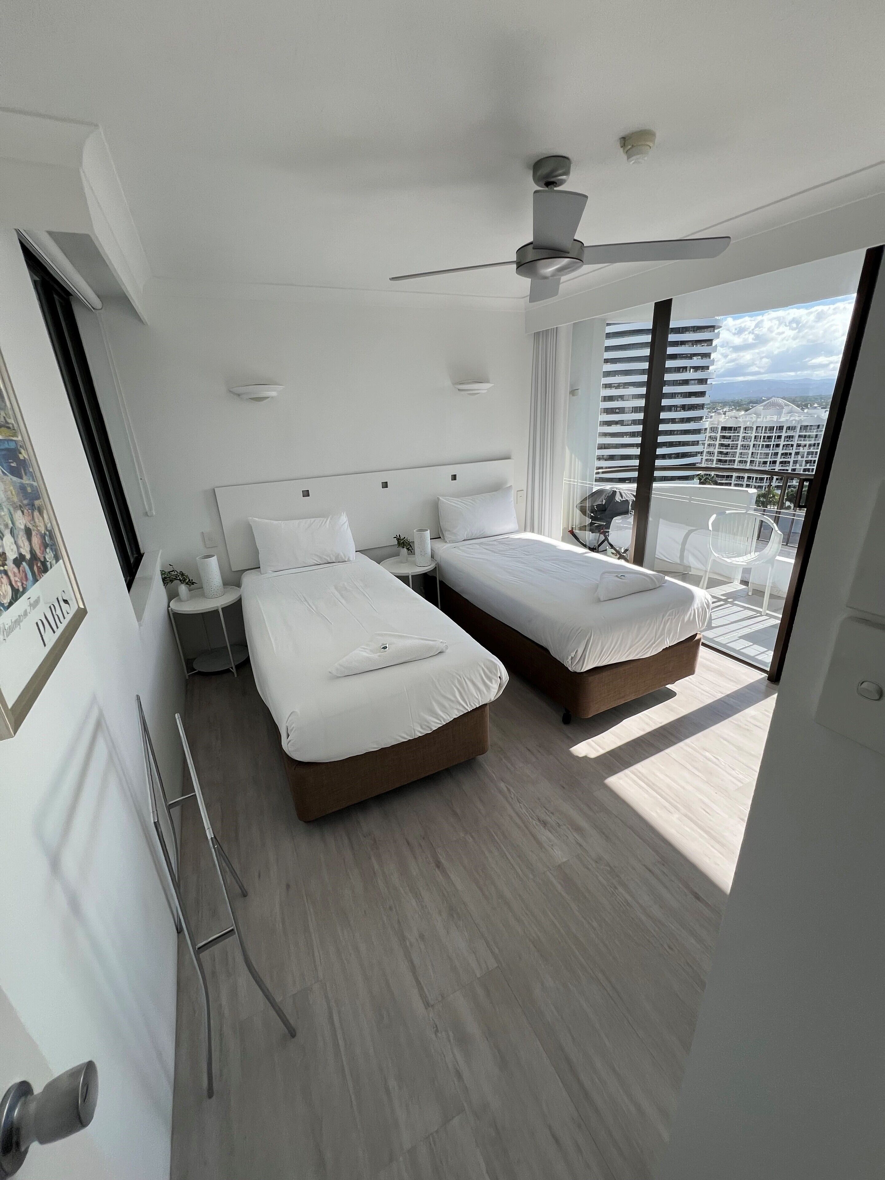 Et moderne hotelværelse med to senge, en loftsvifte, udsigt over byen og en balkon med en stol.