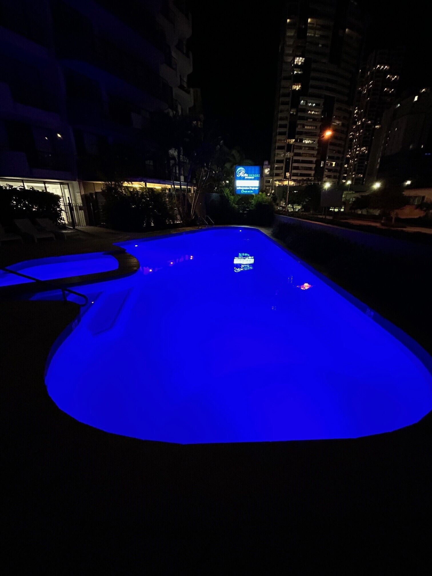 A nighttime view of a swimming pool with blue lighting, surrounded by buildings and a signboard.