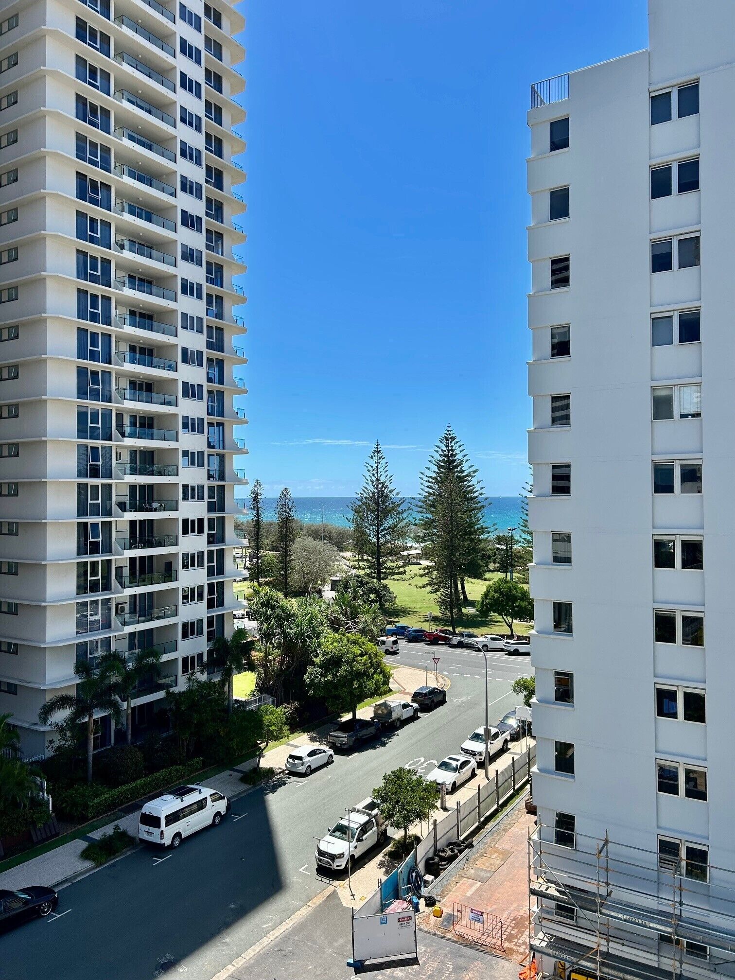 A high-rise building with multiple balconies, a parking lot with cars, and a clear view of the ocean.