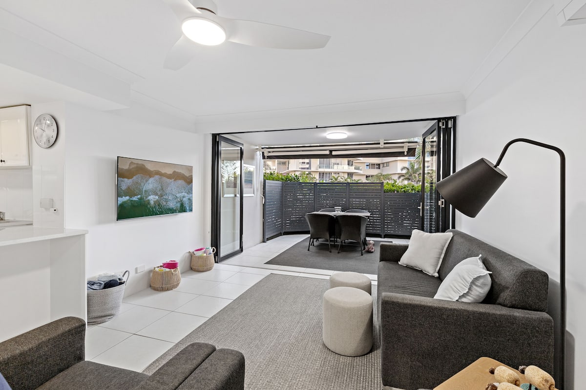 A modern living room with a grey sofa, a coffee table, and a TV mounted on the wall. There is a dining area with a table and chairs visible through a glass door.
