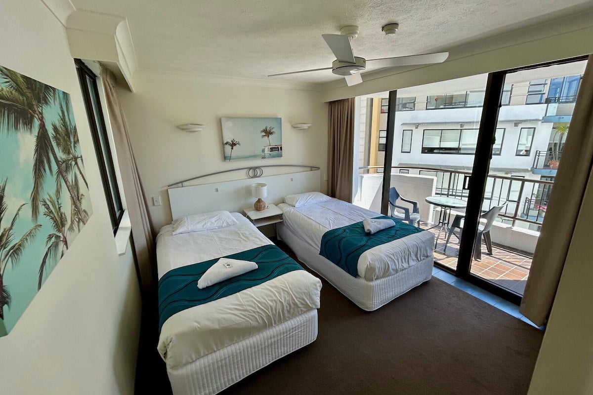 A hotel room with two beds, a ceiling fan, a painting, and a balcony with chairs.