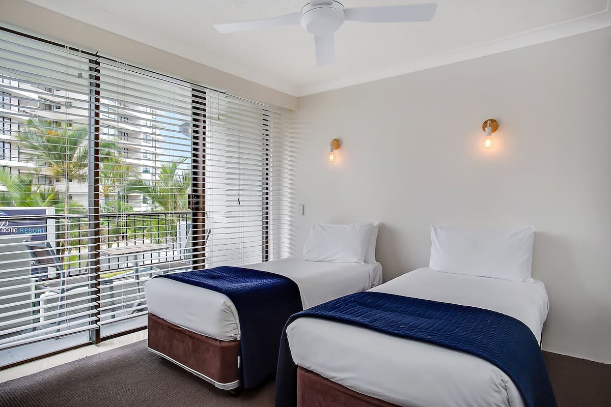 A hotel room with two beds, a ceiling fan, and a view through large windows.