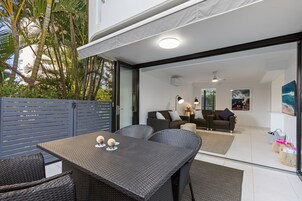 Un área de comedor al aire libre con mobiliario de mimbre y un ventanal de vidrio que da a una sala de estar con sofá y mesa de centro.