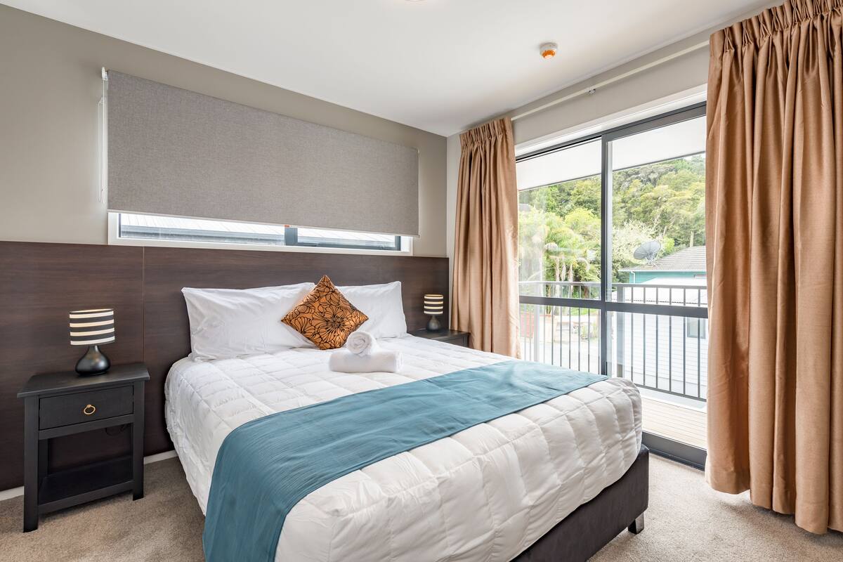 A hotel room with a large bed, a nightstand with a lamp, a window with curtains, and a view of greenery outside.