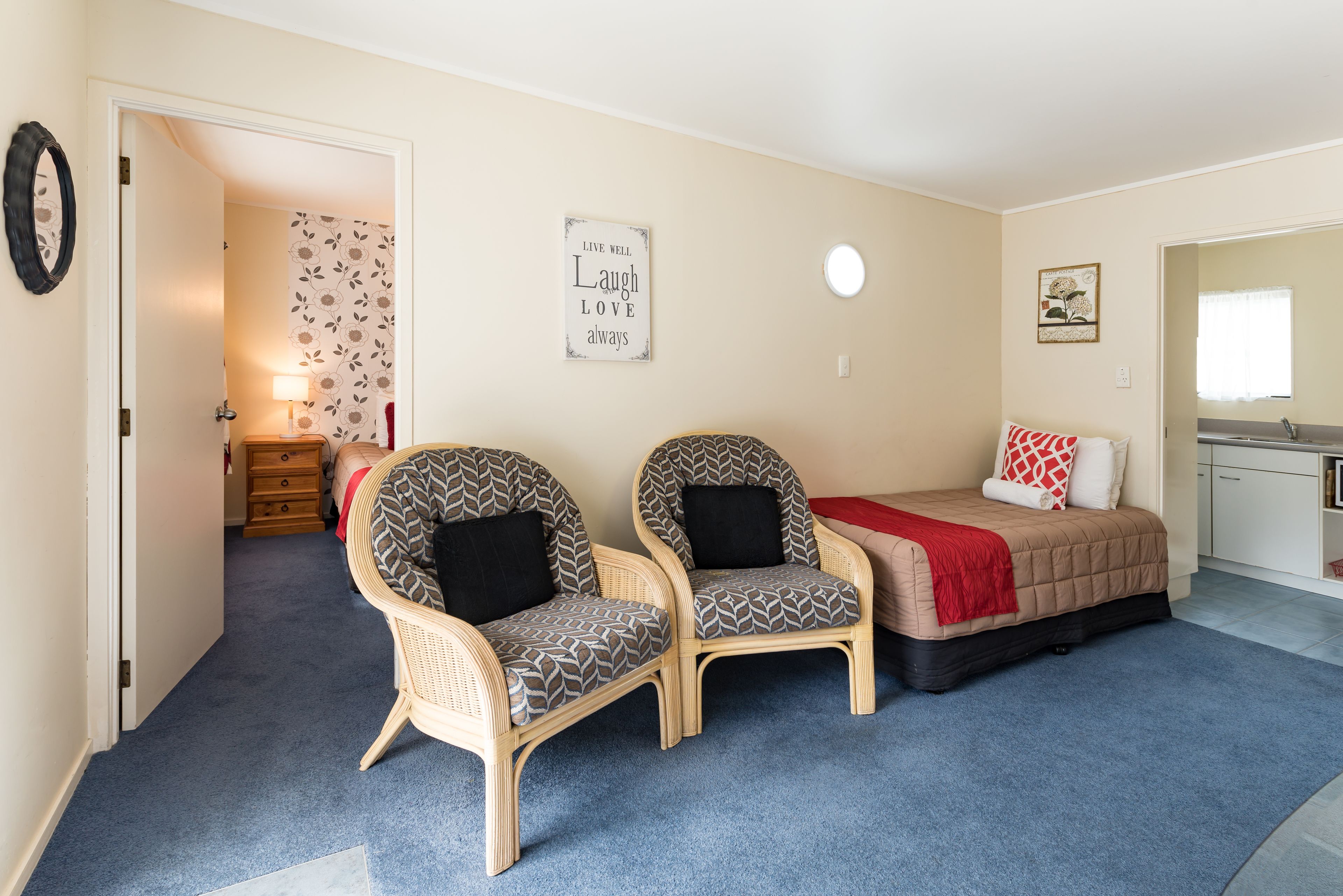 Ein Schlafzimmer mit zwei Korbstühlen, einem Bett mit roter Decke, einer Wand mit der Aufschrift 'Live Well, Laugh Love Always' und einem kleinen Fenster.