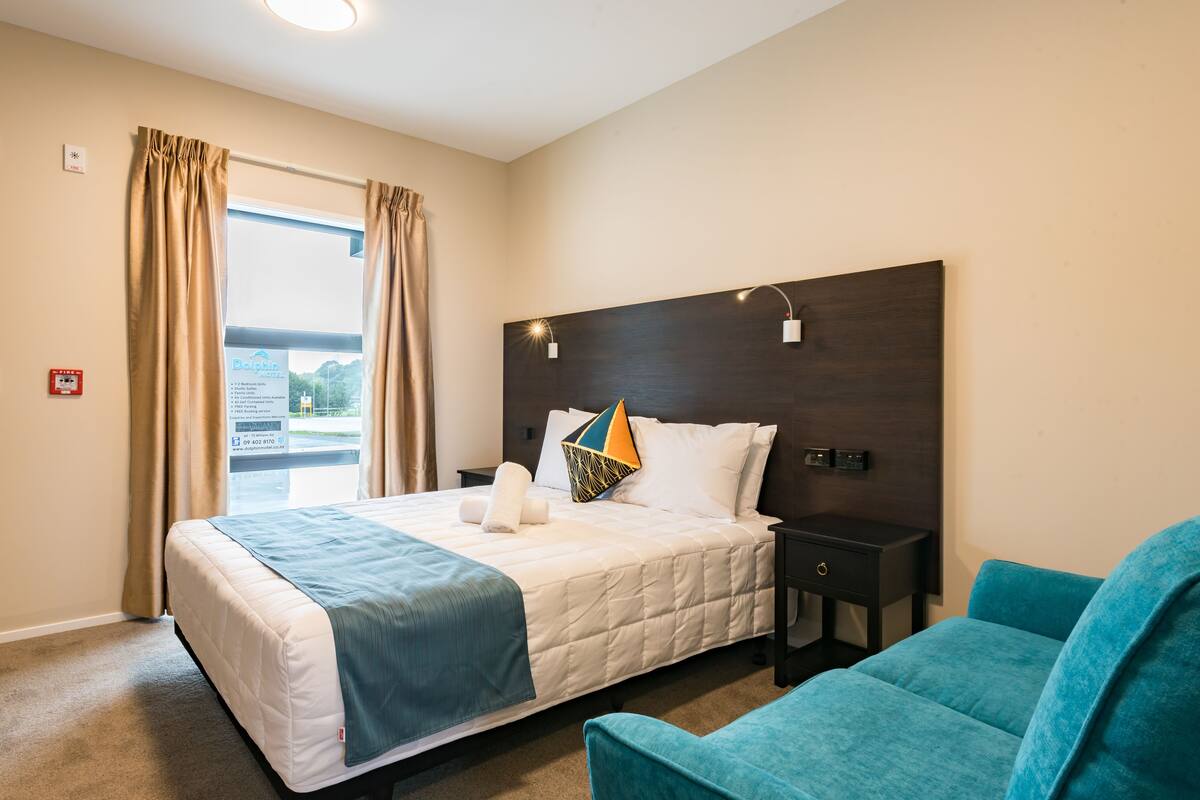 A hotel room with a bed, a nightstand, a chair, a window with curtains, and a sign on the wall.