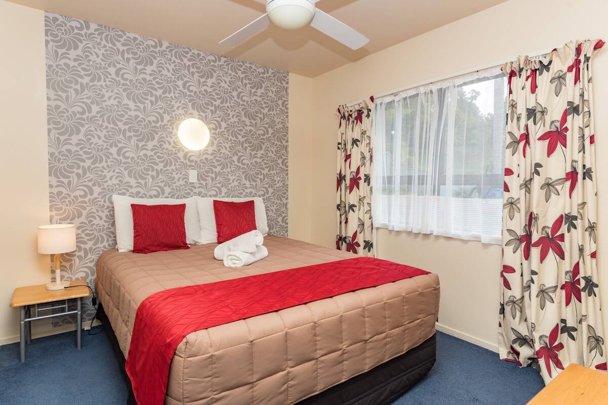 A neatly made bed with a red blanket, a bedside table with a lamp, floral curtains, and a ceiling fan.