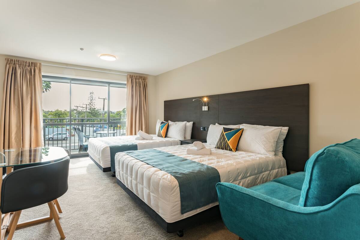 A hotel room with two beds, a glass table, teal chairs, and a balcony with a view of the city.