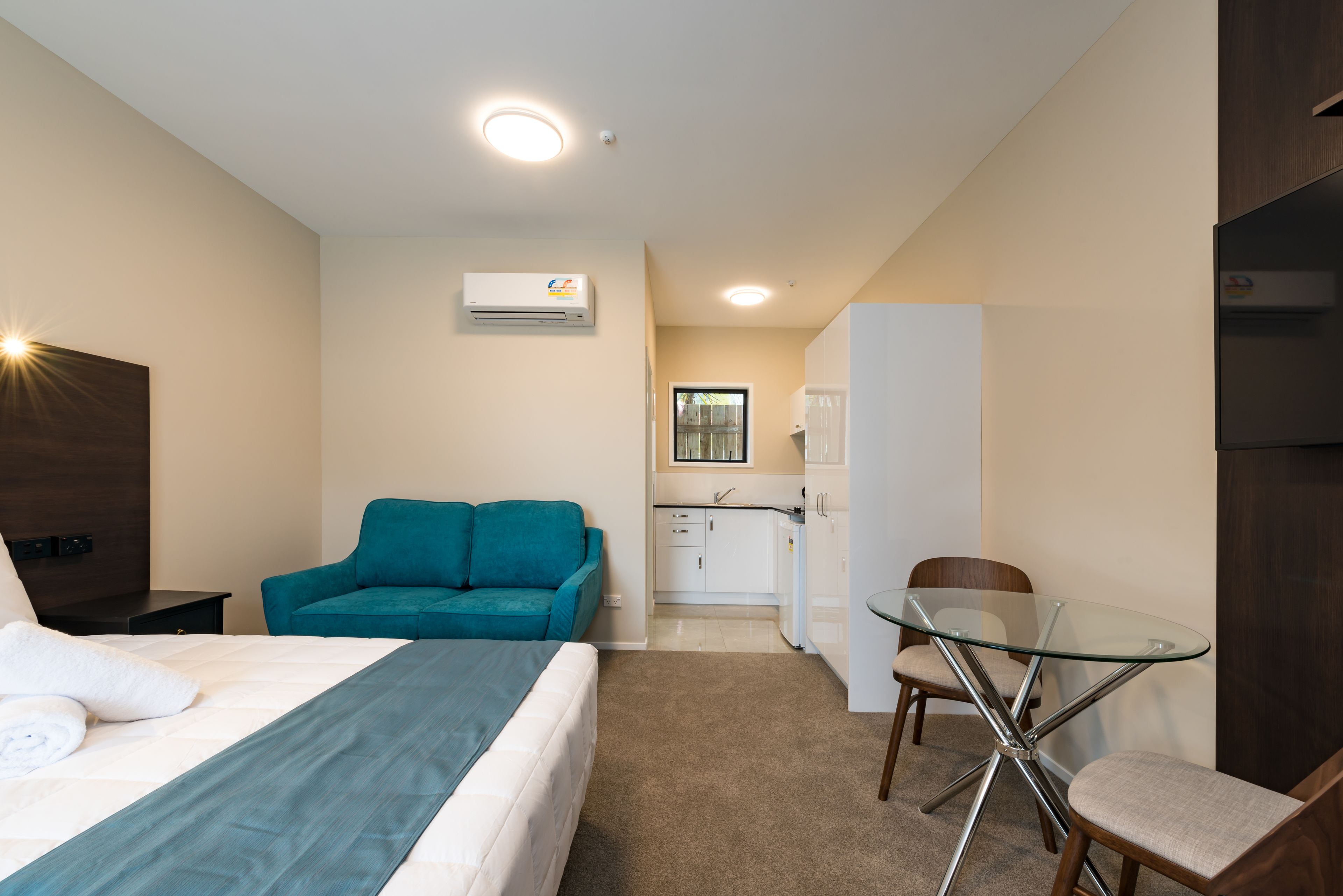 A hotel room with a bed, a teal sofa, a glass table with chairs, a kitchenette, and an air conditioning unit.