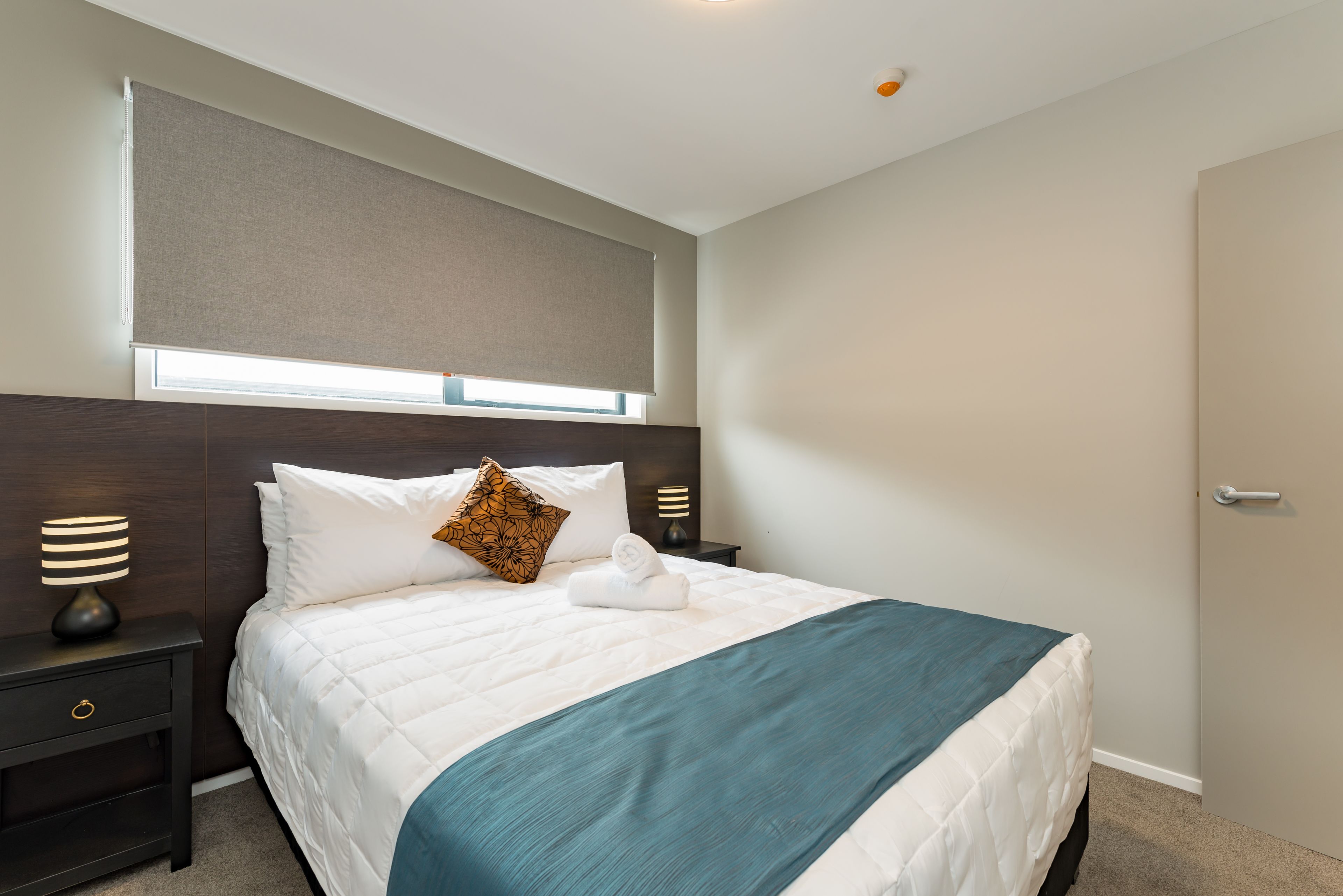 A neatly made bed with a white duvet and a teal throw blanket, a wooden headboard, a nightstand with a lamp, and a window with a beige blind.