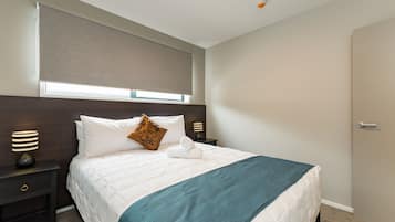 A neatly made bed with a white duvet and a teal throw blanket, a wooden headboard, a nightstand with a lamp, and a window with a beige blind.