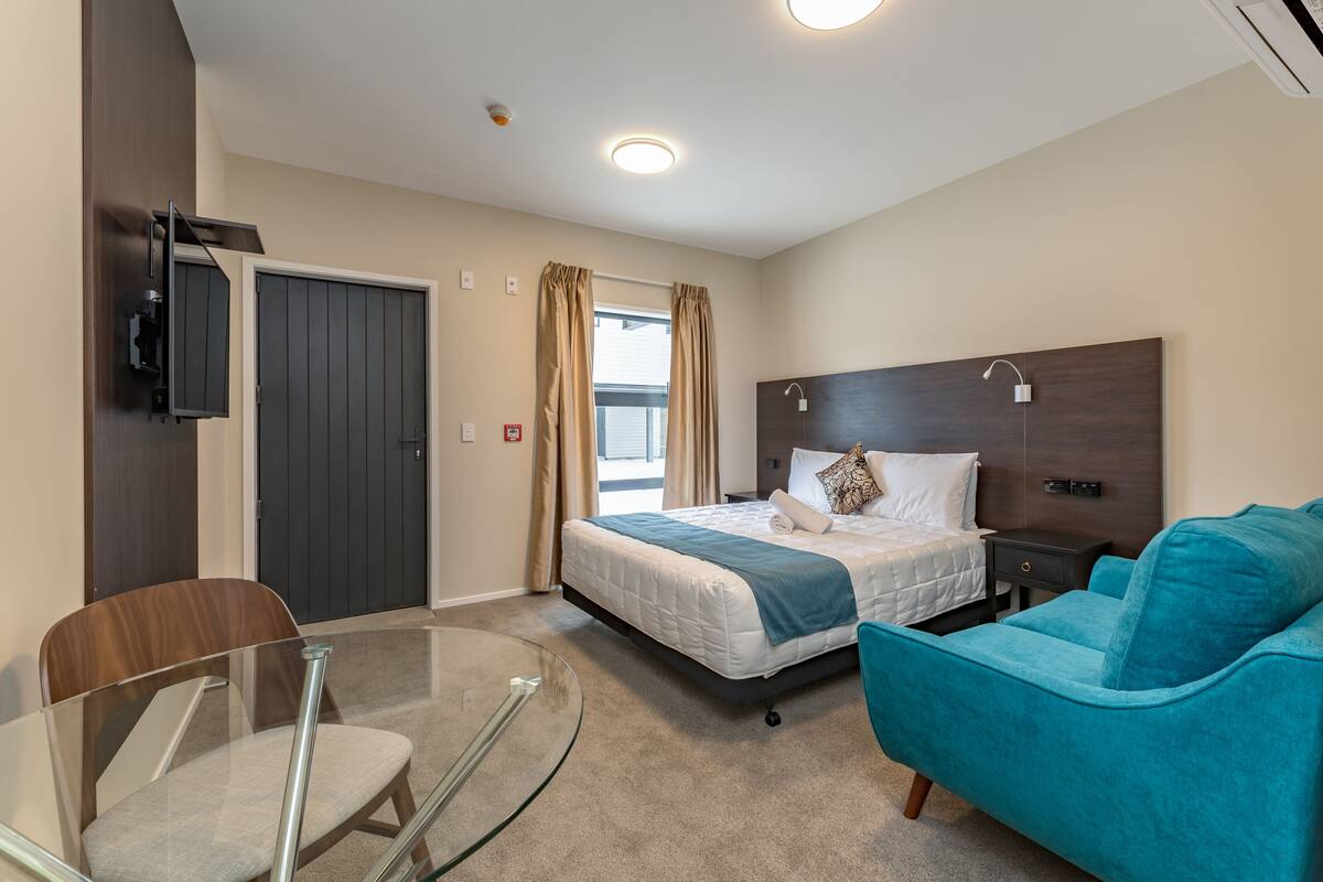 A hotel room with a bed, a glass table, a teal sofa, and a TV mounted on the wall.