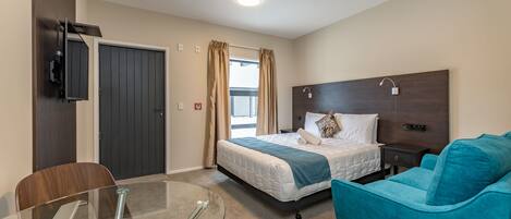 A hotel room with a bed, a glass table, a teal sofa, and a TV mounted on the wall.