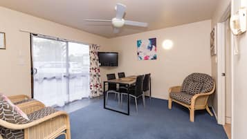 Una habitación con ventilador de techo, televisión, una mesa de comedor con sillas, una silla de mimbre y un gran ventanal con cortinas.