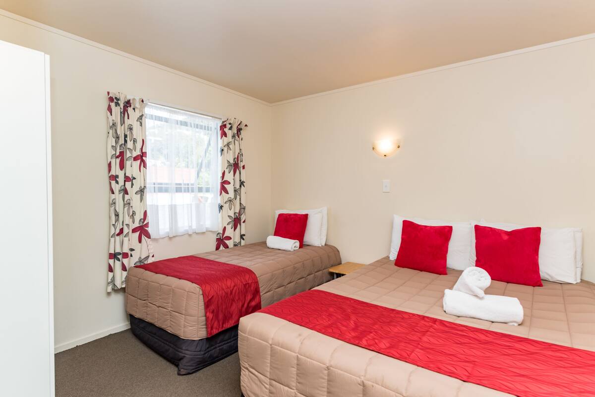 A hotel room with two beds, each with red and white bedding, a window with floral curtains, and a wall-mounted light fixture.