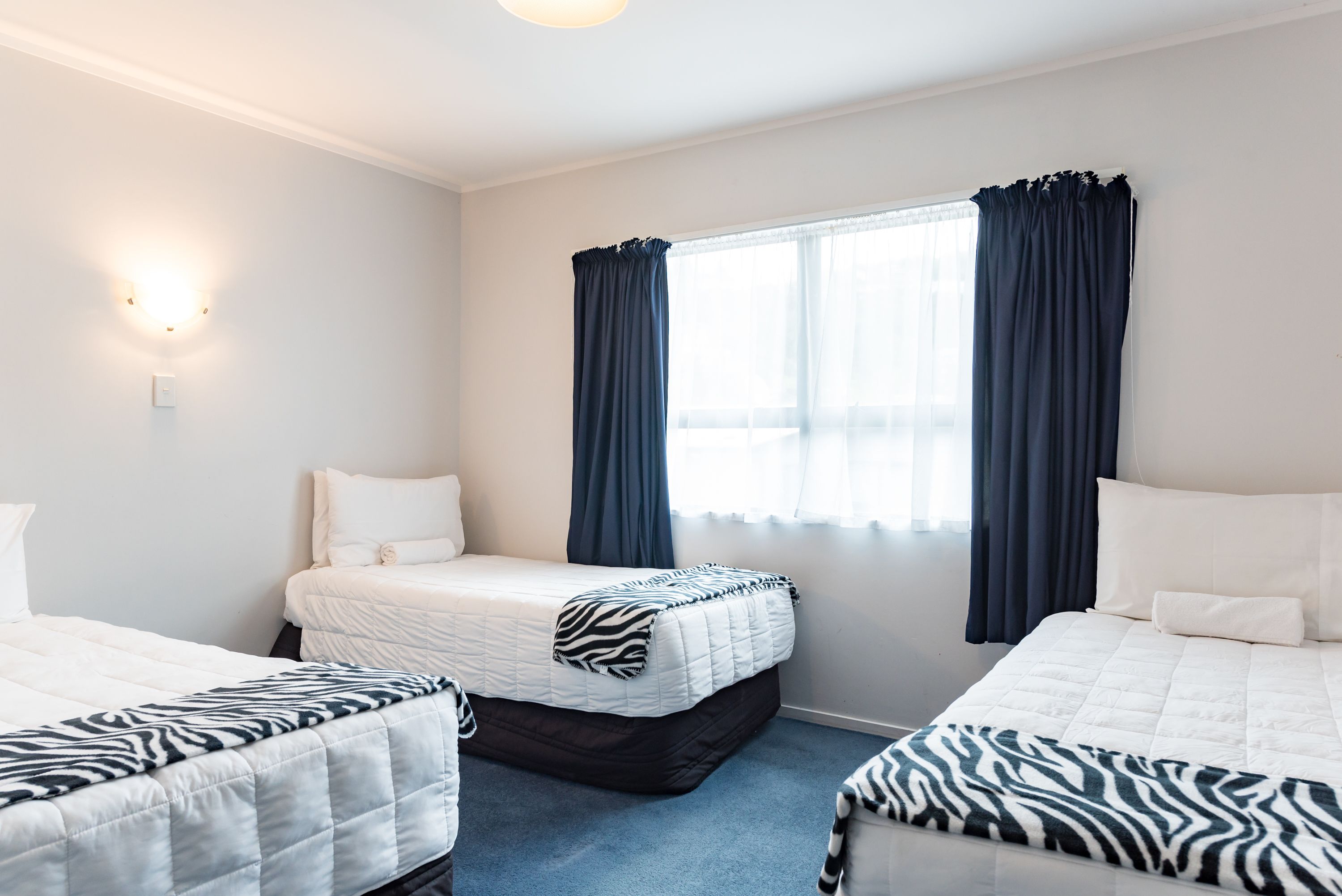 A hotel room with two beds, a large window with curtains, a wall-mounted light, and a carpeted floor.