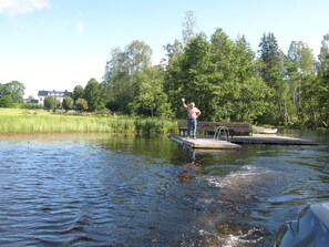 A person standing on a dock extending into a body of water, surrounded by greenery and trees. - Sikfors Herrgård (Hallefors)