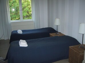 A hotel room with two beds, a window with a view of greenery, a lamp on a wooden nightstand, and a small table with a lamp. - Sikfors Herrgård (Hallefors)