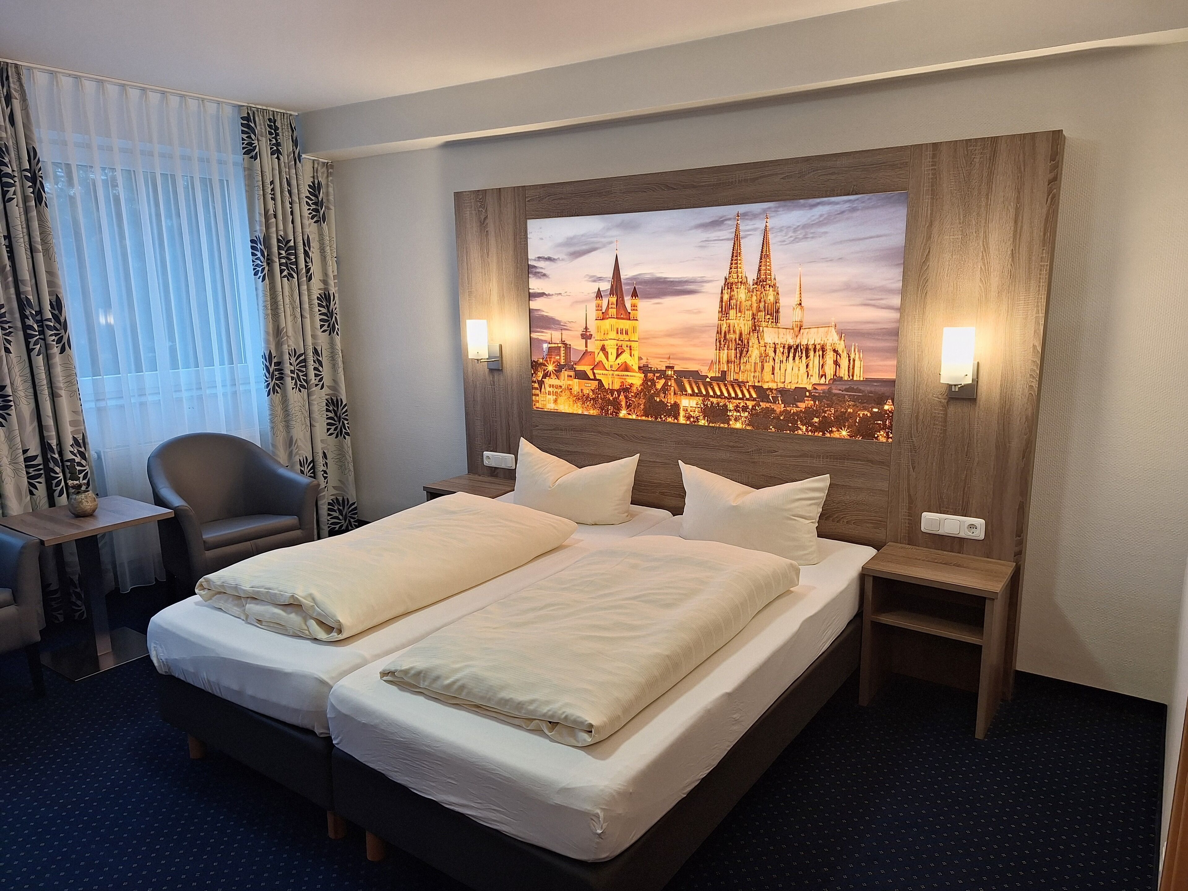 A hotel room with a large bed, two bedside tables, a chair, and a large window with curtains.