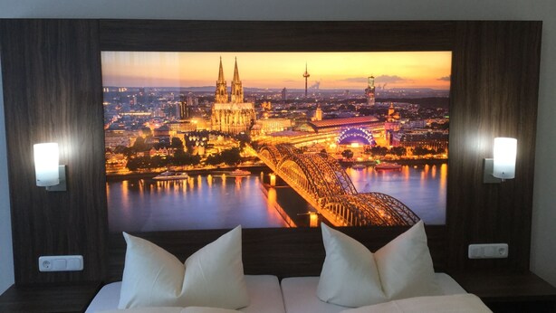 A hotel room with a large wall-mounted TV displaying a cityscape at sunset, a bed with white bedding, and two bedside lamps. - Hotel Am Freischuetz (Huerth)