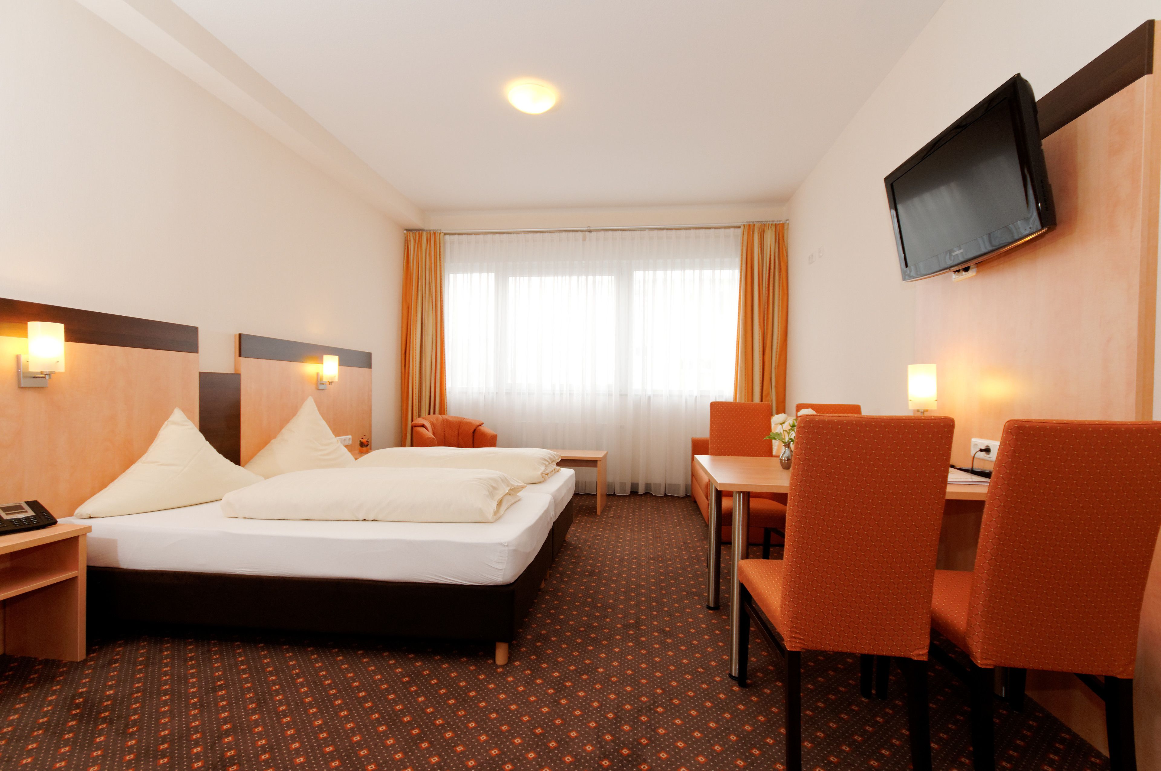 Ein Hotelzimmer mit Bett, Schreibtisch, zwei Stühlen, Fernseher und einem fensterbepflanzten Fenster.
