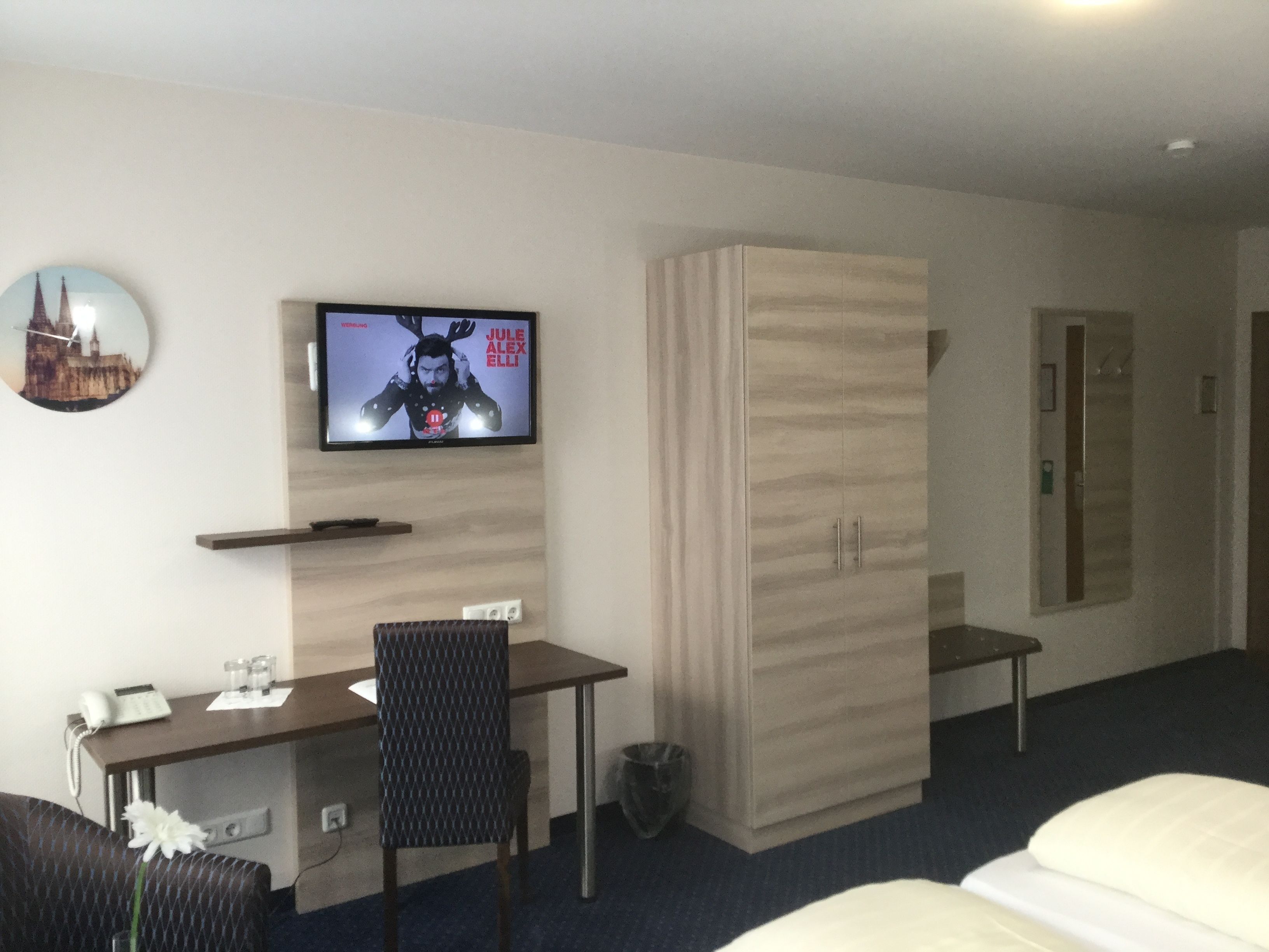 Une chambre d’hôtel comprenant un lit, un bureau avec une chaise, une télévision, une armoire et une petite table avec un téléphone et des verres.