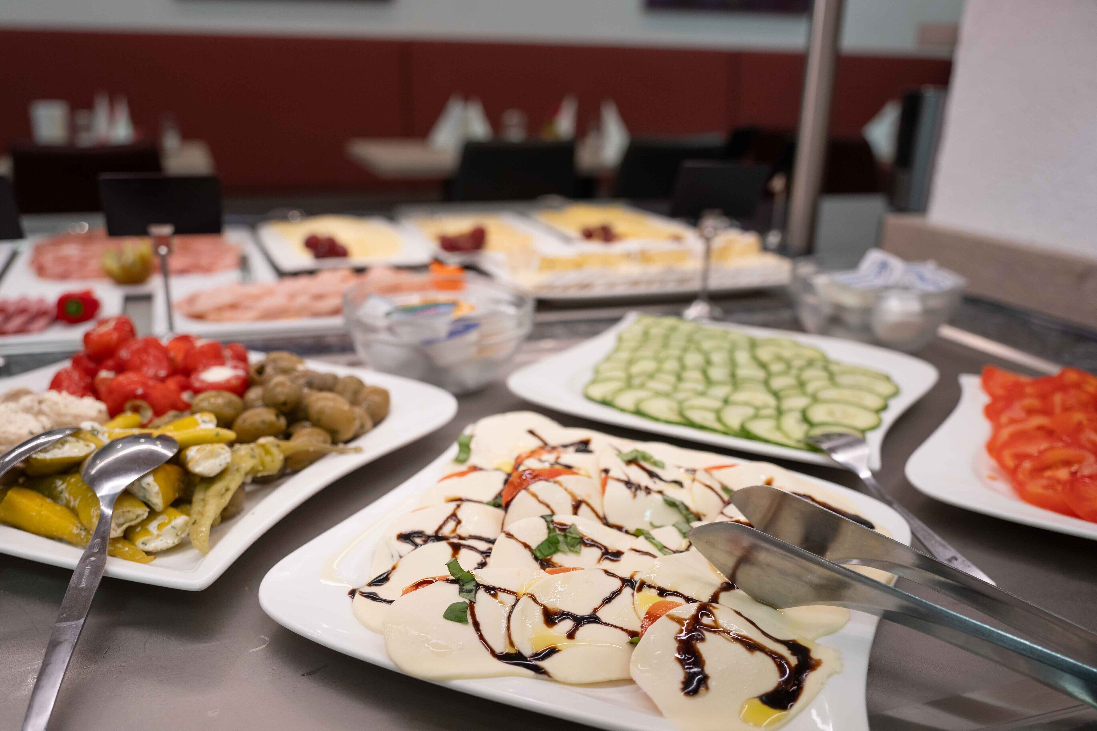A buffet table with various food items including sliced cucumbers, tomatoes, and a dish with white slices drizzled with sauce.