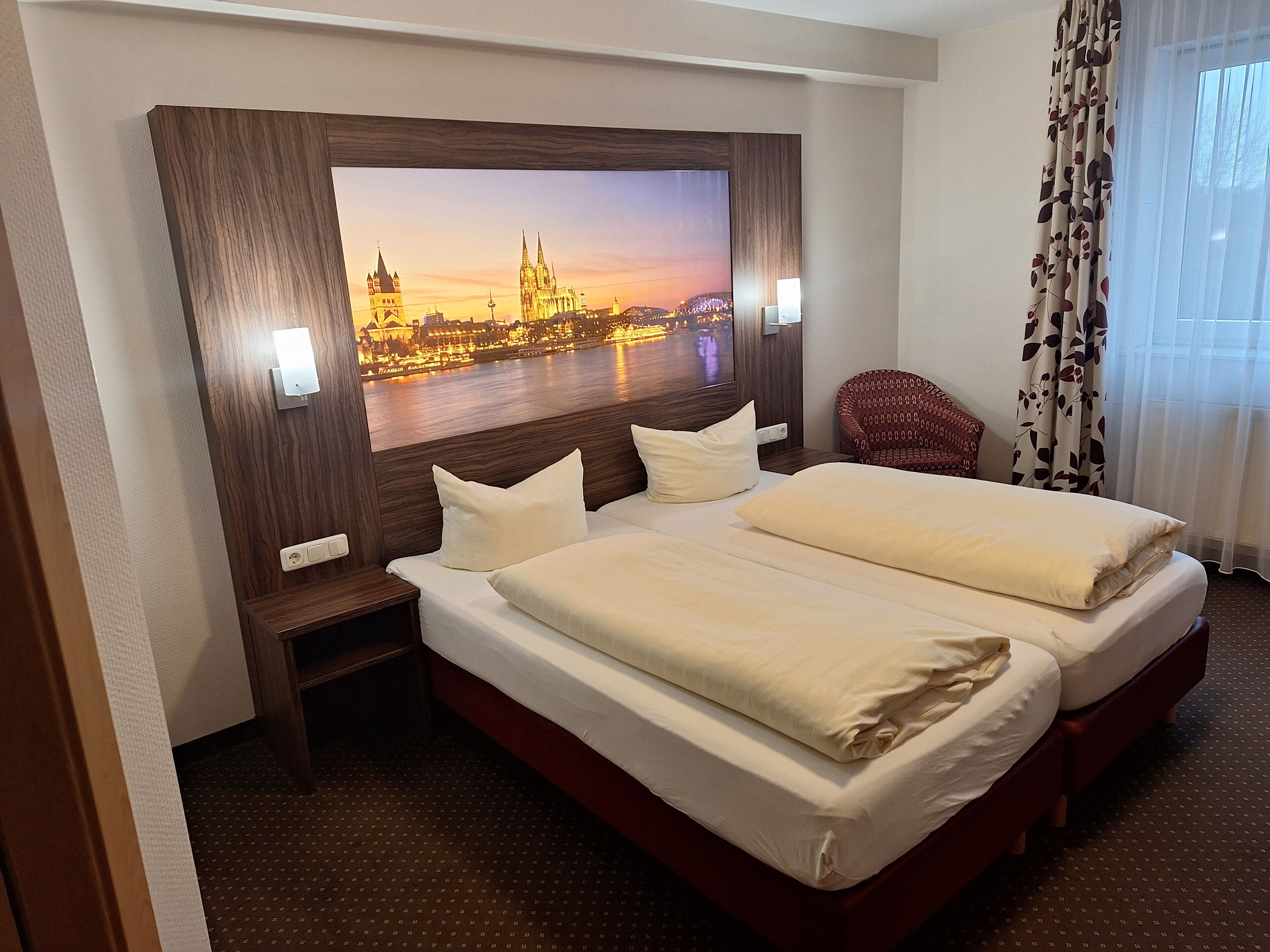 A hotel room with a large bed, a chair, a TV displaying a cityscape, and a window with curtains.