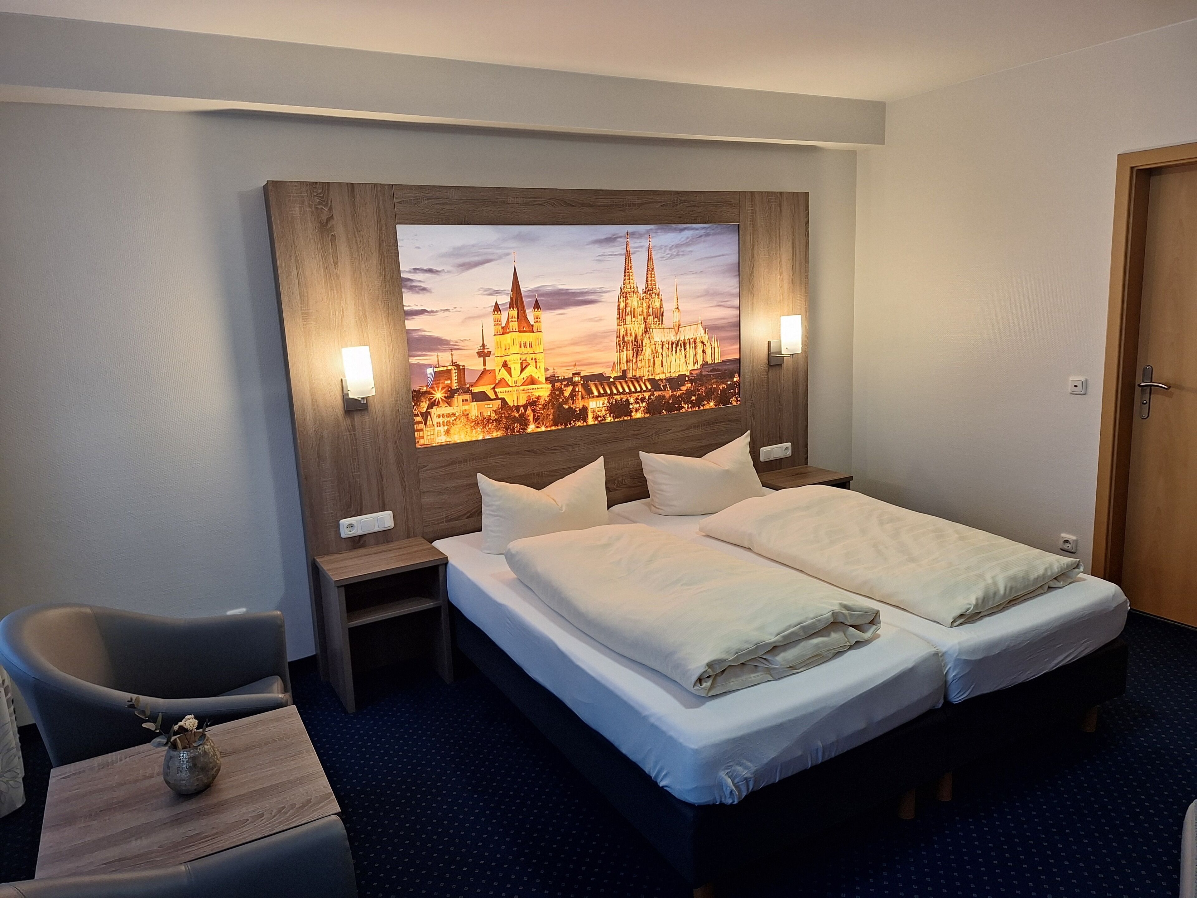 Een hotelkamer met een groot bed, twee nachtkastjes, een stoel en een kunstwerk van een stadsgezicht, bevestigd aan de muur.