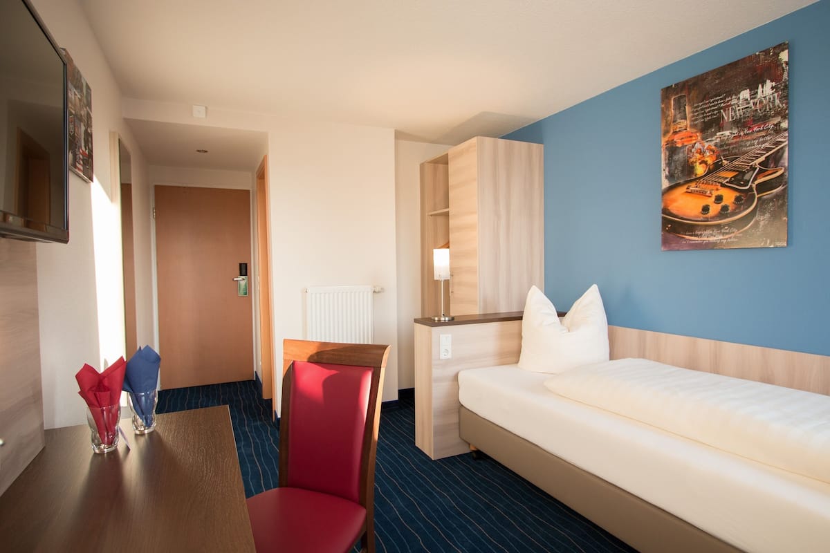 A hotel room with a bed, a desk with a chair, a table with napkins, and a painting on the wall.